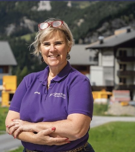 Sele Doris Frau mit Brille und lila Shirt lächelt vor einer Berglandschaft und Gebäuden.